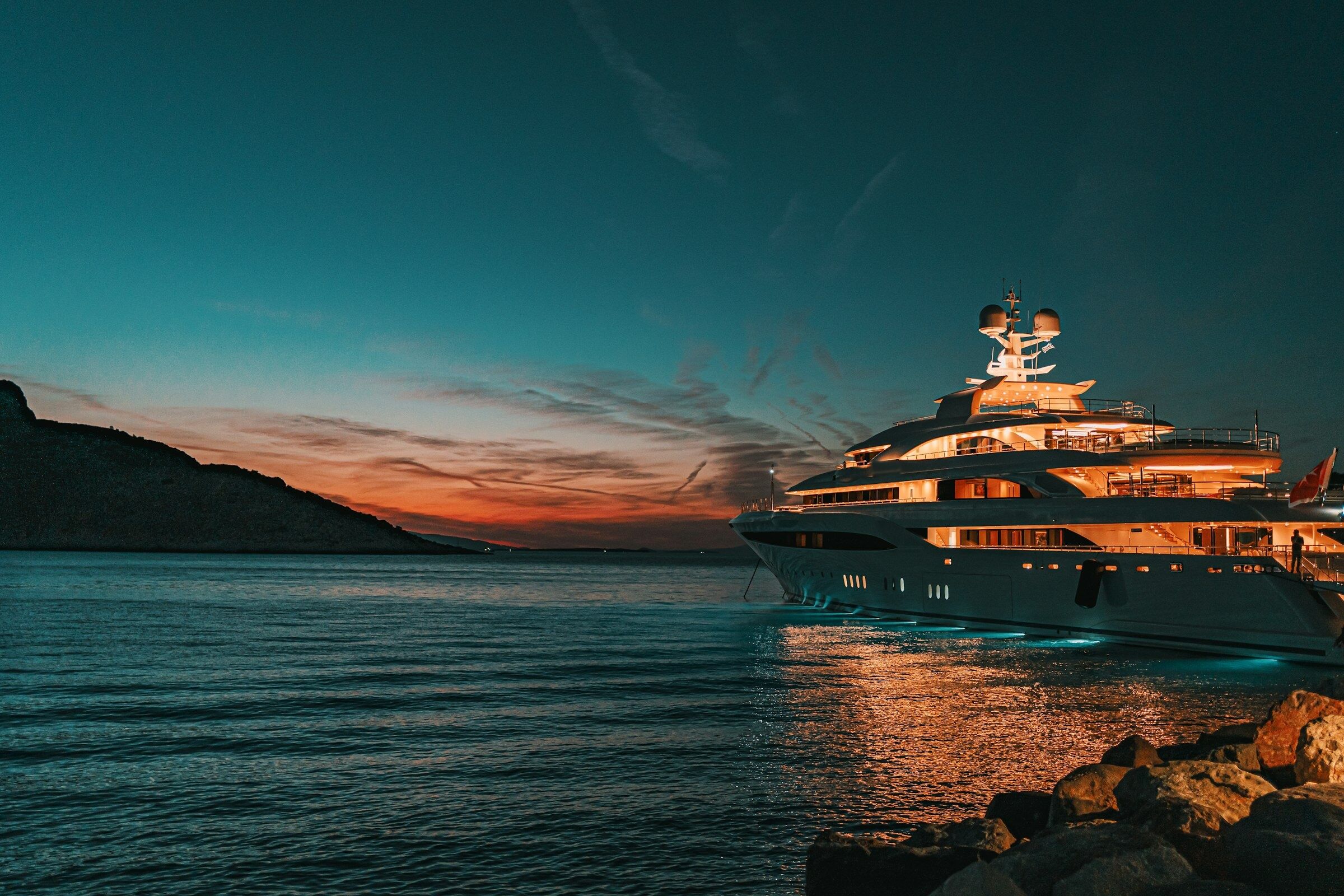 A superyacht lit up at dusk, moored beside a rocky shore
